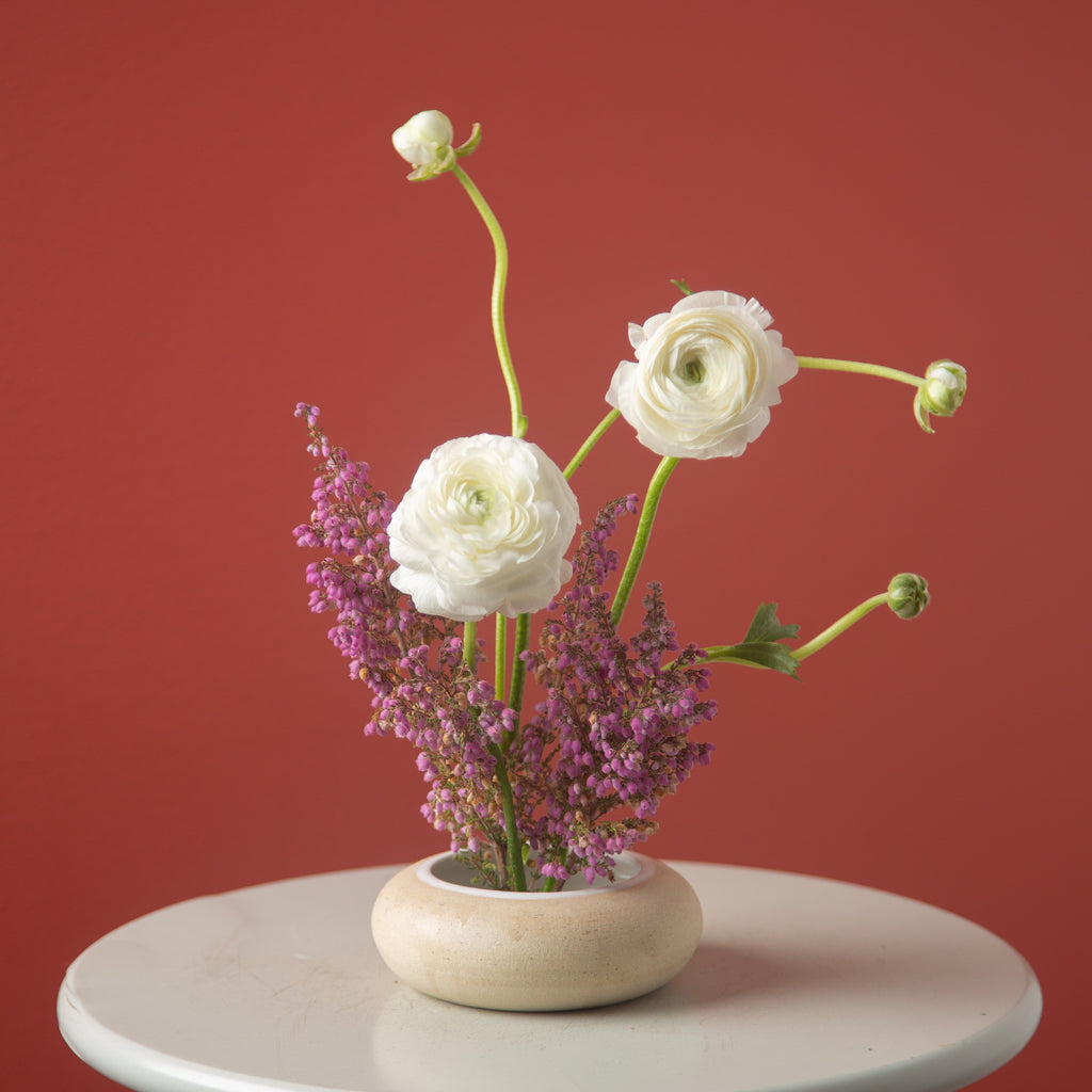 Floral arrangement with white and purple flowers in a wooden vase on a white table against a red background
