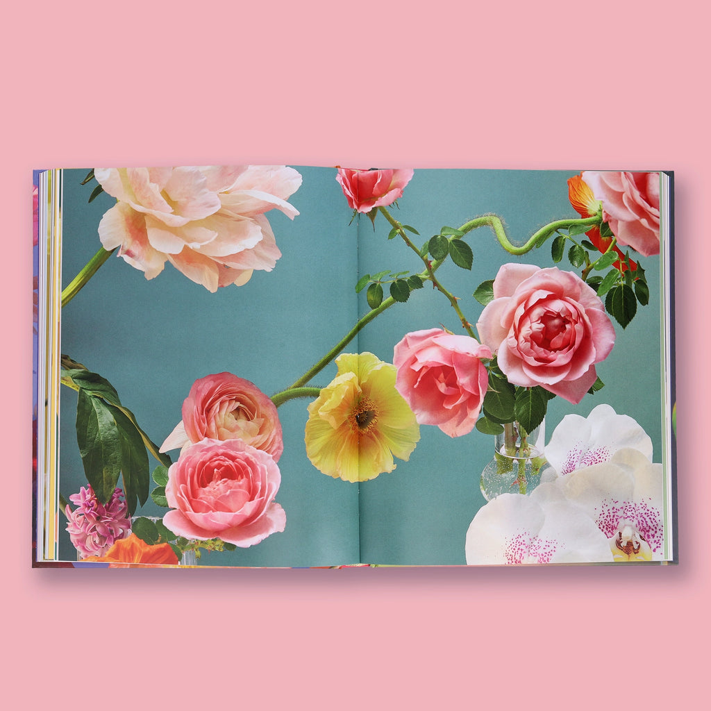 An open book featuring a floral illustration with pink and peach roses, a yellow flower, green leaves, and white orchids on a teal background, set against a solid pink backdrop.